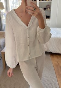 Top alb, texturat, cu un decolteu volan, cu nasturi în față și mâneci tip balon, asortat cu pantaloni de o nuanță deschisă.