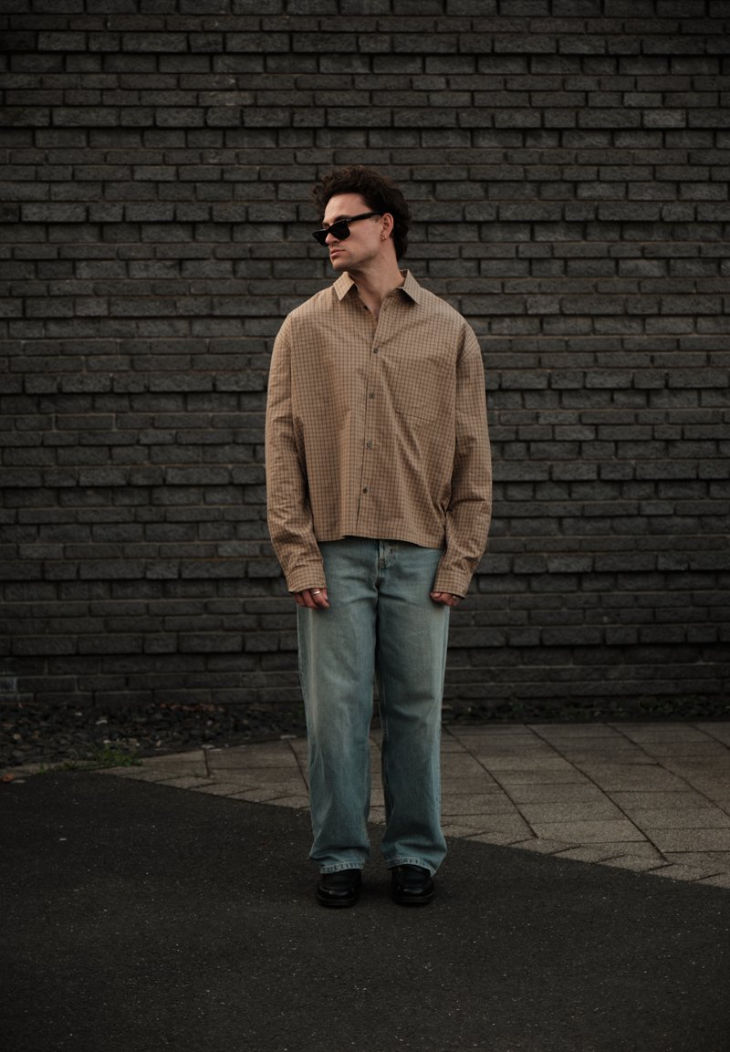 Homme portant une chemise à carreaux beige surdimensionnée, un jean bleu clair, des chaussures noires et des lunettes de soleil, debout devant un mur de briques foncé.