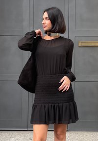 Μαύρο φόρεμα με μακριά μανίκια, διαφανές πάνω μέρος, σμπωχτό μέσον και ριχτό στρίφωμα. Συνδυασμένο με μια μαύρη σουέτ τσάντα και μεταλλικό κολιέ.