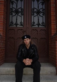 Zwarte leren jas, zwart shirt en zwarte baret gedragen door een persoon die op stenen trappen zit voor sierlijke bruine dubbele deuren.