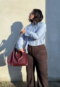 Grande mala de mão burgundy com uma superfície texturizada, combinada com uma camisa de manga longa azul clara e calças castanhas contra uma parede cinza.