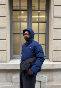 Chaqueta acolchada azul con capucha, con un logo rojo y blanco, llevada sobre una prenda exterior negra acolchada. De pie junto a una ventana grande.