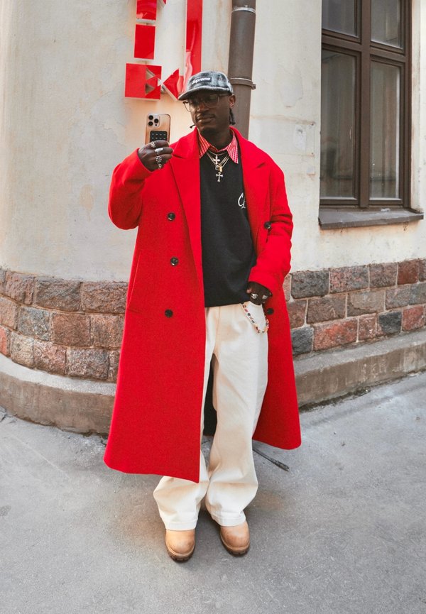 Mann mit rotem Mantel, schwarzem Pullover, weissen Hosen und Mütze macht draussen neben einer Steinmauer und einem Fenster ein Selfie mit dem Smartphone.