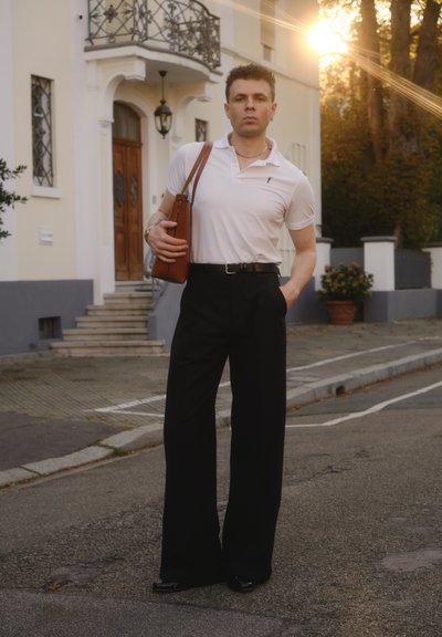 Camisa polo blanca, pantalones anchos negros y bolso marrón de hombro. El entorno presenta arquitectura residencial y un cálido atardecer.