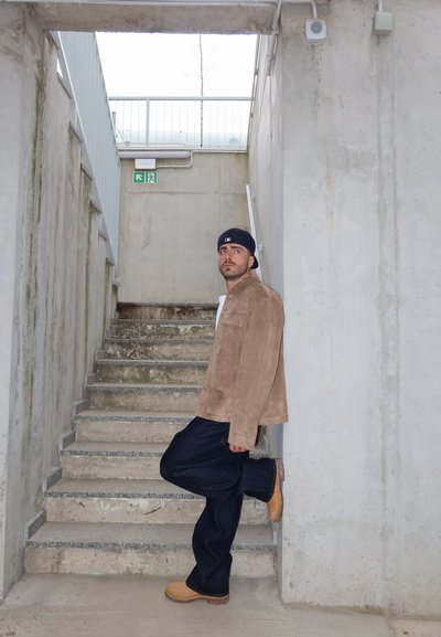 Hombre con gorra, chaqueta marrón, pantalones oscuros y botas color beige apoyado con un pie en una pared de hormigón junto a una escalera en un entorno industrial.