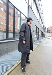 Άνδρας με μαύρο καπέλο, παλτό και παντελόνι στέκεται σε αστικό πεζοδρόμιο και κοιτάζει προς σύγχρονο κτήριο με μεγάλα γυάλινα παράθυρα.