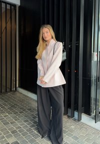 Blazer structuré rose pâle avec un col montant, associé à un pantalon large noir. Tissu lisse et design épuré ; accent du sac bandoulière noir.