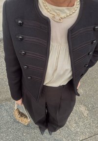 Schwarze Militärjacke mit strukturiertem Design, schwarzen Knöpfen und strukturierten Akzenten über einer cremefarbenen Bluse. Schwarze Hosen mit weitem Bein, perlenbesetzte Handtasche.