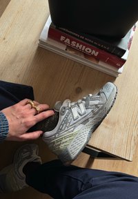Γκρι αθλητικά παπούτσια New Balance με υφή στην επάνω επιφάνεια, ανακλώμενα στοιχεία και καουτσούκ σόλα. Χέρι που ξεκουράζεται πάνω στο παπούτσι, φορώντας πολλούς δακτυλίους.