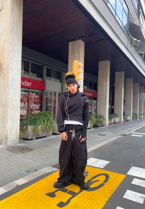 Persona joven con gorro negro y ropa negra de gran tamaño, de pie sobre el símbolo amarillo del carril bici en un entorno urbano.