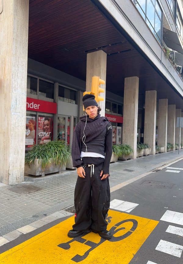 Persona joven con gorro negro y ropa negra de gran tamaño, de pie sobre el símbolo amarillo del carril bici en un entorno urbano.