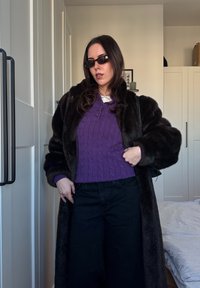 Manteau en fausse fourrure marron sur un pull violet en maille torsadée, associé à un pantalon noir à jambes larges. Les accessoires comprennent des lunettes de soleil foncées et un sac à main.