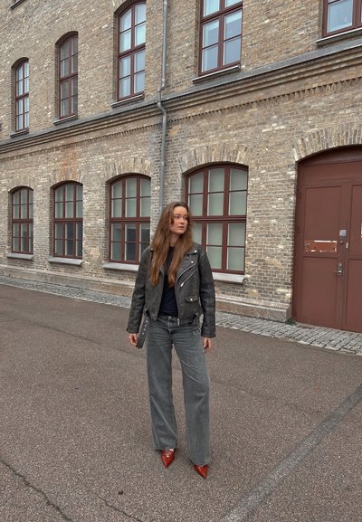 Chaqueta de cuero negro de motorista, camiseta azul marino, jeans anchos grises y tacones rojos puntiagudos. Pelo largo castaño, de pie contra una pared de ladrillo con ventanas.