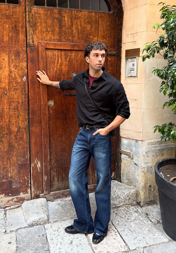 Joven con pelo rizado, vestido con una camisa negra y vaqueros azules, está de pie con una mano apoyada en una gran puerta de madera en una calle empedrada.