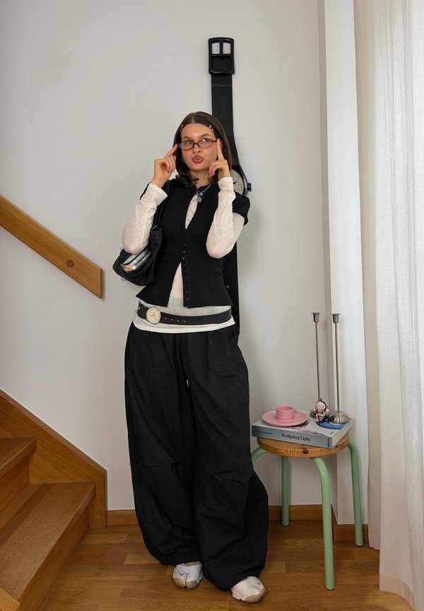 Joven mujer con pantalones negros de gran tamaño y chaleco posa en el interior cerca de unas escaleras, con un reloj de pared negro de gran tamaño y una pequeña mesa con libros cerca.