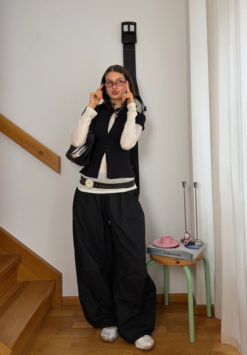 Joven mujer con pantalones negros de gran tamaño y chaleco posa en el interior cerca de unas escaleras, con un reloj de pared negro de gran tamaño y una pequeña mesa con libros cerca.