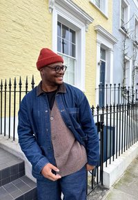 Jachetă din denim peste un cardigan maro tricotat, asortată cu o căciulă roșie. Ochelari de soare închiși la culoare, stând lângă o casă galbenă cu balustrade negre.