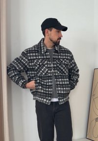 Casaco de flanela em padrão xadrez preto e branco, com uma gola grande, dois bolsos no peito e fecho de correr frontal, usado sobre um suéter cinzento e jeans pretos.