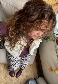 Krémová prešívaná vesta cez vínový top, spárovaná s nohavicami s leopardím vzorom. Tmavé vínové čižmy, štruktúrovaná tkanina a viditeľná vnútorná zeleň.