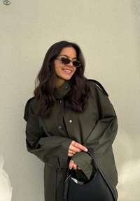 Ελαιοπράσινο μακρύ σακάκι με ψηλό γιακά και κουμπιά, συνδυασμένο με μαύρη τσάντα υφής και γυαλιά ηλίου με λεοπάρ σχέδιο.