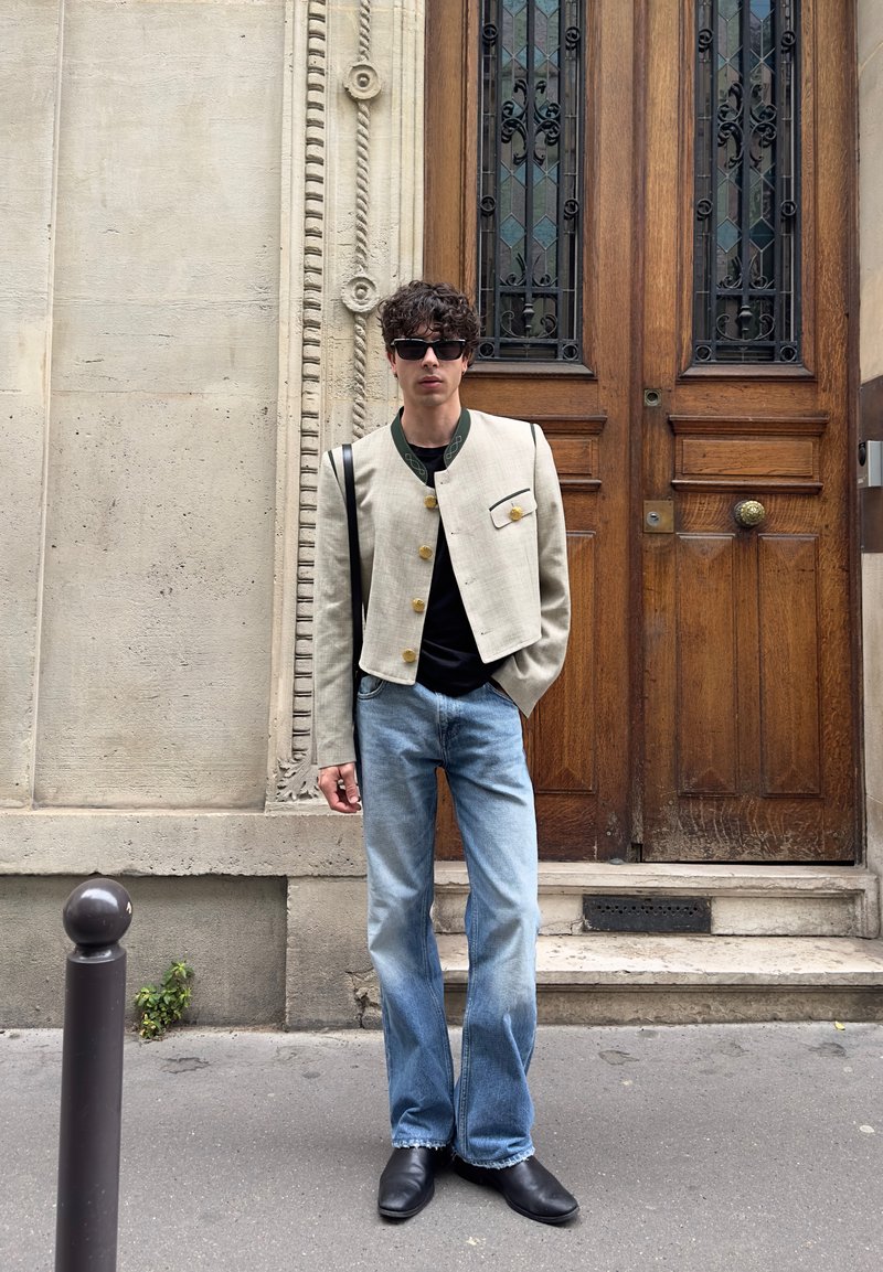 Jeune homme portant des lunettes de soleil, un blazer beige court, une chemise noire, un jean bleu et des chaussures noires, debout sur un trottoir en ville devant des portes en bois.