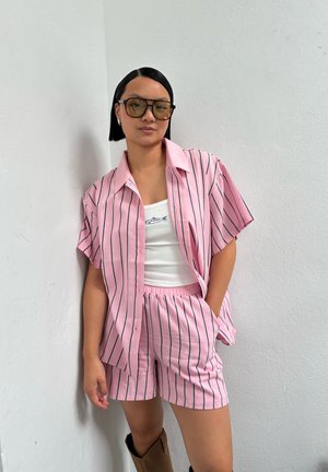 Camisa de manga corta a rayas rosas y pantalones cortos a juego, hechos de tela ligera. Lleva una camiseta sin mangas blanca debajo. Los accesorios incluyen gafas de sol de gran tamaño.