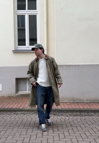 Jeune homme portant un trench-coat vert, un pull blanc en maille torsadée, un jean bleu et une casquette, debout sur une rue pavée près d'un bâtiment beige avec une fenêtre.
