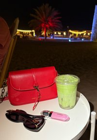 Sarkanas ādas crossbody soma ar ķēdes siksnu, caurspīdīga glāze ar zaļu dzērienu, melni saulesbrilles un rozā kosmētikas produkts uz baltas galda.