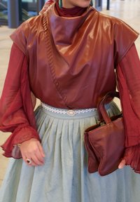 Personne portant un haut en cuir bordeaux sur un chemisier rouge transparent, une jupe plissée bleu clair avec une ceinture en perles, tenant un sac à main en cuir bordeaux.