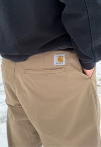 Personne portant un pantalon Carhartt beige avec un sweat-shirt noir, mains dans les poches, debout dehors sur une surface enneigée.