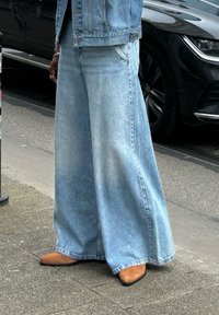 Vaqueros de mezclilla de pierna ancha en un tono azul claro con una textura deslavada, combinados con zapatos de punta afilada en color marrón claro. El conjunto presenta una silueta casual y holgada.