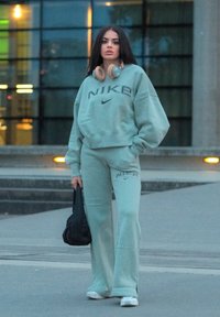 Lys grønn Nike sweatshirt og matchende bukser med logo, kombinert med hvite sneakers og hodetelefoner, stående på fortau.