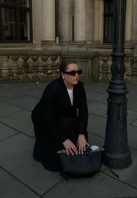 Juodas odinis rankinukas su trumpomis rankenėlėmis atidarytas guli ant pilkos dangos. Žmogus juodame paltuke nusilenkia prie gatvių lemp�ų, taisydamas rankinuką.