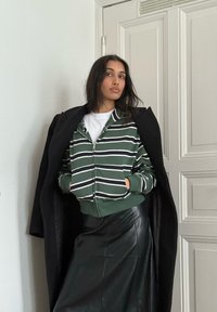 Femme portant un sweat à capuche zippé vert et blanc rayé par-dessus un t-shirt blanc, une jupe en cuir noire et un manteau noir posé sur les épaules à l'intérieur près d'une porte.