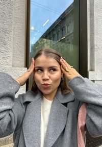 Grau-wolliger Mantel mit übergroßen Ärmeln, darunter ein cremefarbener Pullover und eine pinkfarbene, geflochtene Handtasche. Der Schmuck umfasst Ringe und Creolen.