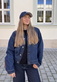 Traper jakna opuštenog kroja, tamnoplave boje, metalni dugmadi, uparen s crnim puloverom i odgovarajućim tamnimfarmerkama. Crna kapa upotpunjuje izgled.