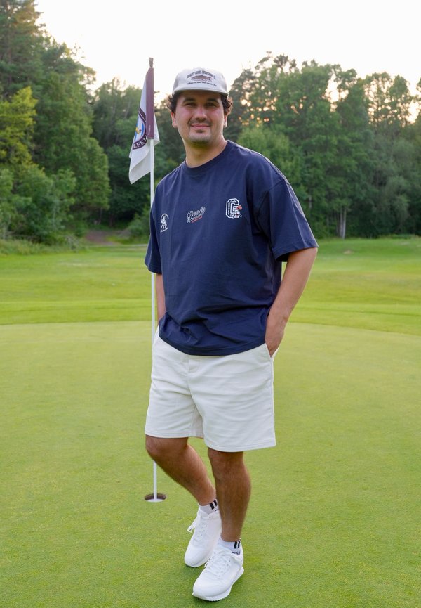 Homme en chemise bleu marine, short blanc et casquette, debout sur le green près du trou avec drapeau, entouré d'arbres en plein jour.