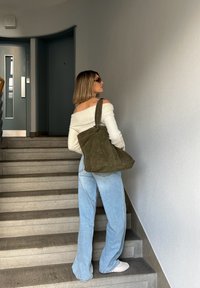 Zeleno semiš torba z velikim, ohlapnim oblikom, enim trakom in subtilnimi šivi, nosi jo na stopnicah v kombinaciji z svetlo modrimi širokimi jeans hlačami.