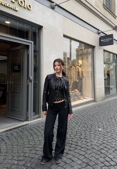 Joven mujer con chaqueta de cuero negra y blusa de lunares de pie en una calle empedrada frente a la tienda Marc O’Polo, con maniquíes en la vitrina.