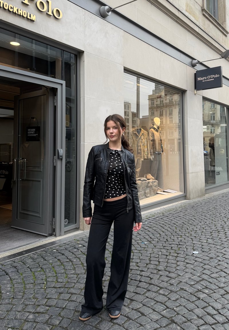 Jauna sieviete melnā ādas jaku un punktainu blūzi stāv uz bruģētas ielas pie Marc O’Polo veikala ar manekinām logu izstādē.