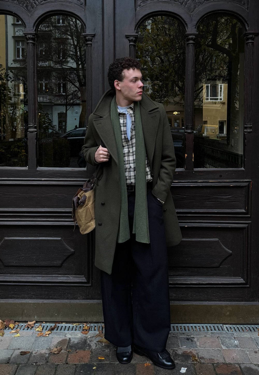 Płaszcz oliwkowo-zielony na koszuli w kratę, w połączeniu z szerokimi czarnymi spodniami. Dodatki to jasnozielony szalik oraz jasnobrązowa torba listonoszka.