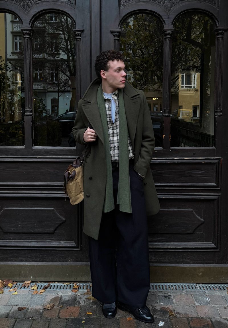 Palton verde măsliniu peste o cămașă în carouri, asortat cu pantaloni largi negri. Accesorii includ o eșarfă verde deschis și o geantă de umăr maro deschis.