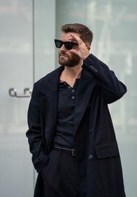 Černý vlněný kabát s širokým límcem, nošený přes černý henley. Osoba má na sobě černé sluneční br�ýle, krátké vousy a vlasy upravené do pořádku.