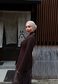 Ung kvinde i beige tørklæde og mørkebrun blazer står sidelæns nær træsløjfer og stofgardin med sort kalligrafi.