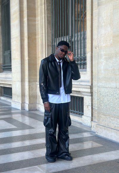 Chaqueta bomber de cuero negro, camisa blanca, pantalones cargo negros con bolsillos y zapatos negros. Gafas de sol estrechas completan el conjunto.