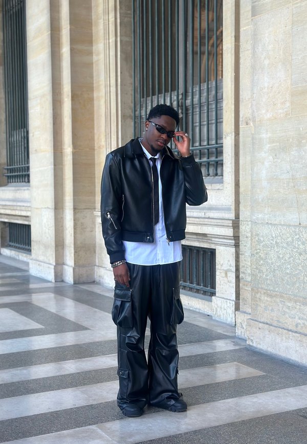 Giacca bomber in pelle nera, camicia bianca, pantaloni cargo neri con tasche e scarpe nere. Occhiali da sole slim completano il look.