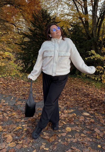 Chaqueta de forro polar cremosa y texturizada con una franja plateada, pantalones anchos negros y zapatillas negras. Grandes gafas de sol y bolso completan el look.