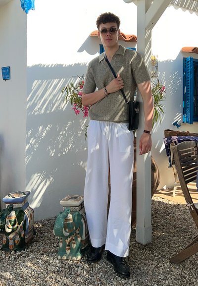 Polo de manga corta de punto en beige claro, combinado con pantalones blancos de cordón. Bolso cruzado negro y zapatos oscuros. Fondo decorativo de plantas.