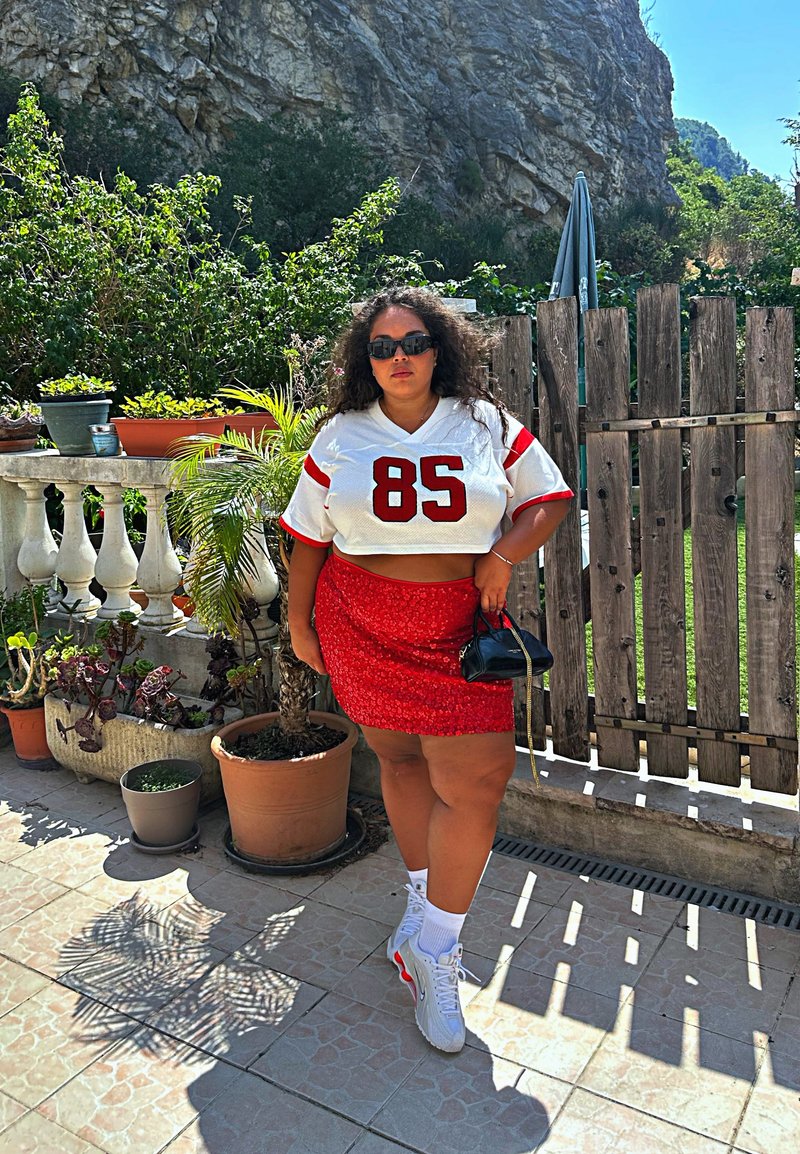 T-shirt blanche cropped avec des accents rouges et "85," jupe à sequins rouges, sac à main noir, baskets blanches avec des détails rouges, dans un cadre de jardin.