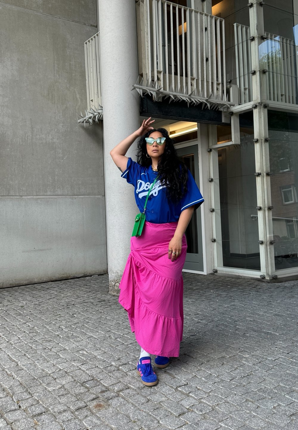 Blue baseball jersey with white lettering, pink tiered maxi skirt, green crossbody bag, and blue trainers. Textured background with stone paving.